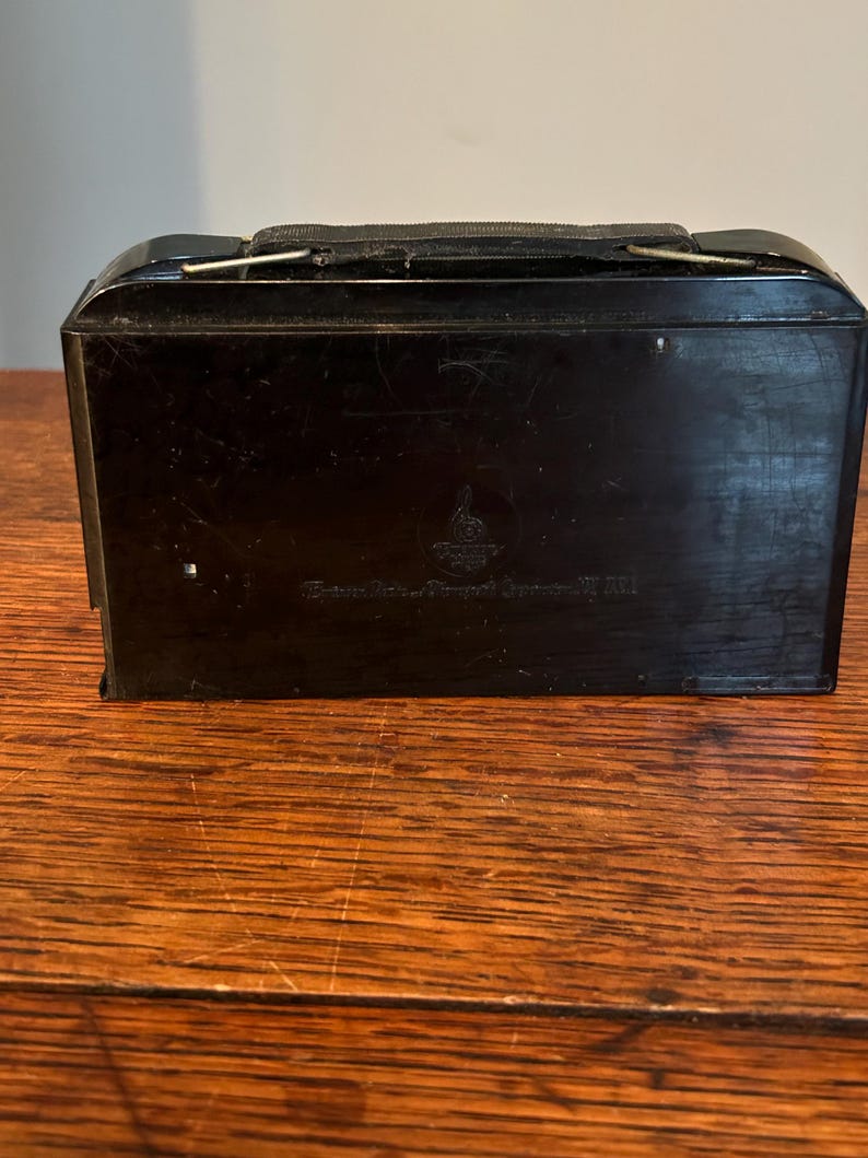 May include: A black, rectangular case with a handle and a clasp. The case has a glossy finish and appears to be made of a hard material. The text on the case is blurry.