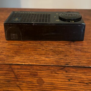 May include: A vintage black radio with a speaker grill and a tuning dial. The dial has numbers 6, 7, 10, and 15. The radio is rectangular and sits on a wooden surface.