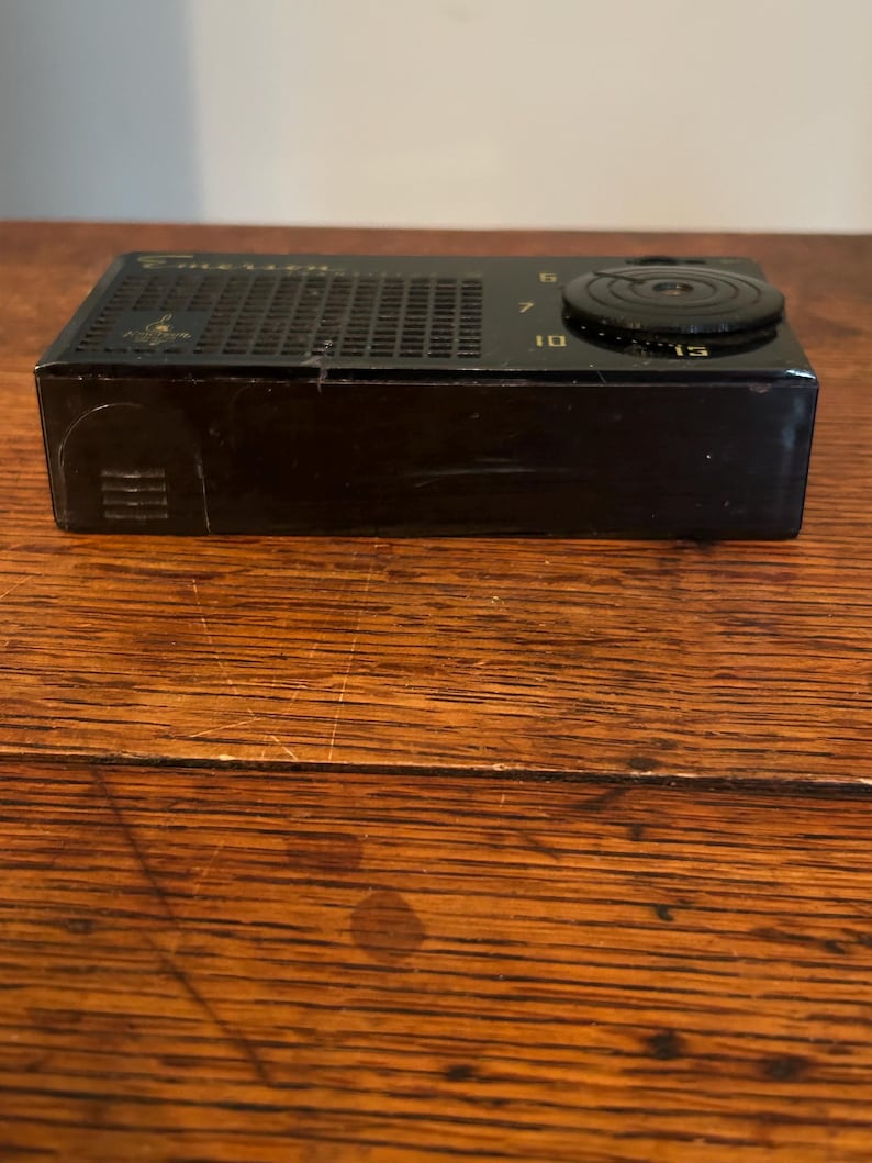 May include: A vintage black radio with a speaker grill and a tuning dial. The dial has numbers 6, 7, 10, and 15. The radio is rectangular and sits on a wooden surface.