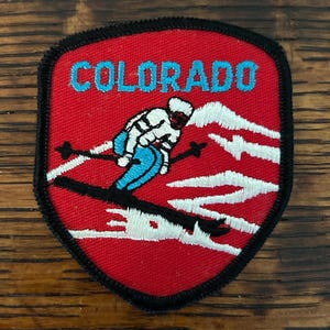 May include: Embroidered patch with a red background, black border, and the word "COLORADO" in turquoise. A skier in white and blue skis down a mountain. The patch is a shield shape.
