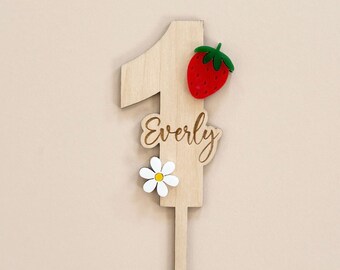 May include: Wooden number one cake topper with the name "Everly" engraved on it. The topper is decorated with a red strawberry and a white daisy.