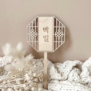 May include: A wooden sign with a traditional Korean design and the text "백일" in Korean. The sign is on a white knitted surface.