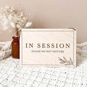 May include: A wooden sign with the text "IN SESSION PLEASE DO NOT DISTURB" in brown lettering. The sign has a small, engraved leaf design in the bottom right corner.