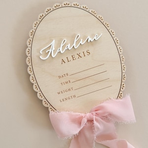 Puede incluir: Un letrero ovalado de madera para anuncio de nacimiento con borde festoneado, que presenta el nombre Adaline Alexis en texto blanco y marr&oacute;n. El letrero incluye l&iacute;neas para fecha, hora, peso y longitud, decorado con una cinta rosa deshilachada.