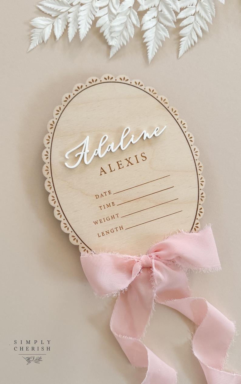 Puede incluir: Un letrero ovalado de madera para anuncio de nacimiento con borde festoneado, que presenta el nombre Adaline Alexis en texto blanco y marr&oacute;n. El letrero incluye l&iacute;neas para fecha, hora, peso y longitud, decorado con una cinta rosa deshilachada.