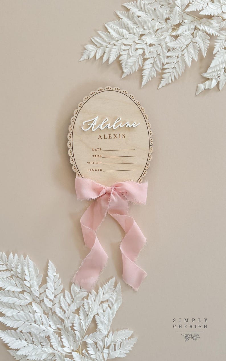 May include: Oval wooden baby announcement plaque with scalloped edge, featuring the names 'Adaline Alexis' in raised white lettering.  Below the names are blank lines for date, time, weight, and length.  A pale pink ribbon is tied below the plaque. The plaque is displayed against a neutral beige background with white ferns.