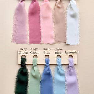 Puede incluir: Dos tablas de madera que sostienen diez cintas de tela con bordes deshilachados en varios colores. La fila superior presenta Dusty Lilac, Mauve, Pink, Nude y White. La fila inferior muestra las etiquetas Deep Green, Sage Green, Dusty Blue, Light Blue y Lavender.