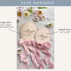 Puede incluir: Dos placas ovaladas de madera con bordes festoneados, grabadas con el nombre Margot Mae. Las placas tienen largas cintas de seda rosa empolvado atadas en la base. El tama&ntilde;o grande mide 13,46 cm por 17,15 cm y el peque&ntilde;o 11,43 cm por 14,61 cm.