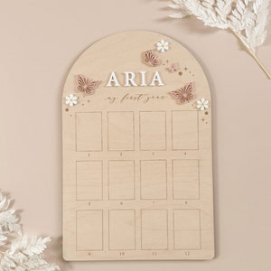 Puede incluir: Un gr&aacute;fico de crecimiento de madera con un fondo marr&oacute;n claro y un arco blanco. El gr&aacute;fico est&aacute; decorado con mariposas y flores. El nombre "ARIA" est&aacute; escrito en letras blancas en la parte superior del gr&aacute;fico. El texto "my first year" est&aacute; escrito en cursiva debajo del nombre. El gr&aacute;fico tiene 12 cuadrados para cada mes del a&ntilde;o.