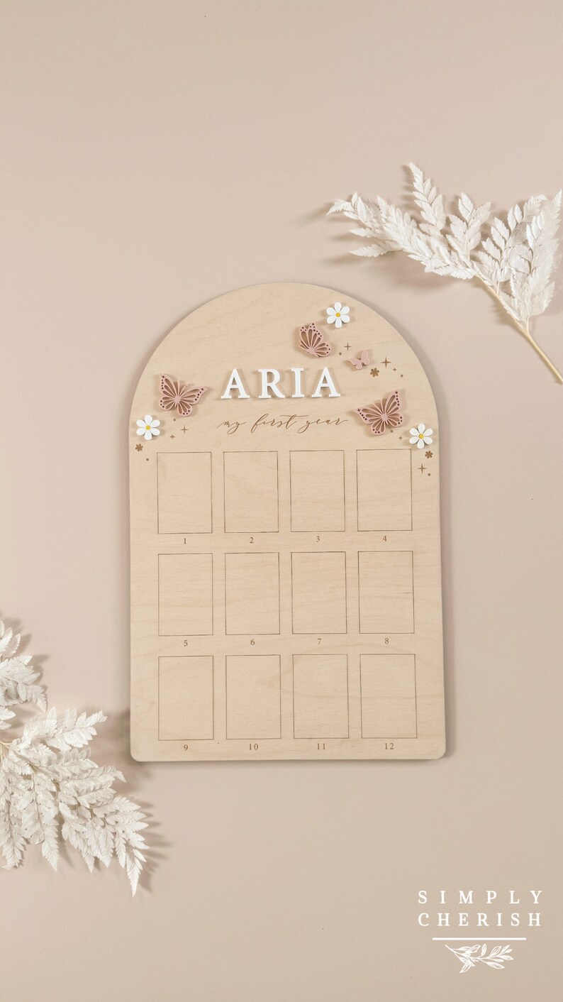 Puede incluir: Un gr&aacute;fico de crecimiento de madera con un fondo marr&oacute;n claro y un arco blanco. El gr&aacute;fico est&aacute; decorado con mariposas y flores. El nombre "ARIA" est&aacute; escrito en letras blancas en la parte superior del gr&aacute;fico. El texto "my first year" est&aacute; escrito en cursiva debajo del nombre. El gr&aacute;fico tiene 12 cuadrados para cada mes del a&ntilde;o.
