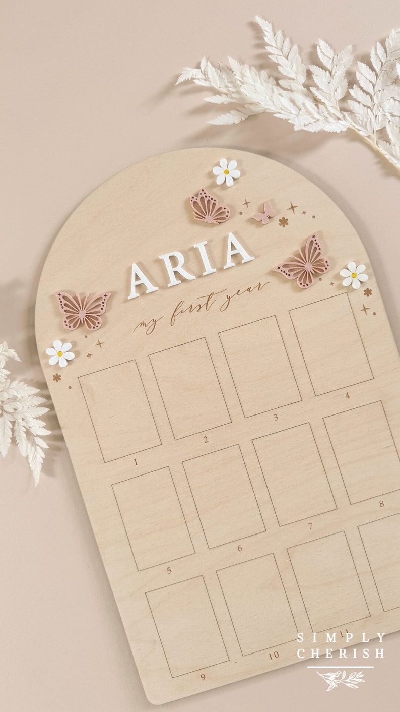Puede incluir: Un gr&aacute;fico de crecimiento de madera en forma de arco con el nombre "ARIA" y el texto "mi primer a&ntilde;o" en cursiva. El gr&aacute;fico tiene 10 espacios en blanco para fotos, numerados del 1 al 10. El gr&aacute;fico est&aacute; decorado con mariposas rosas y flores blancas.