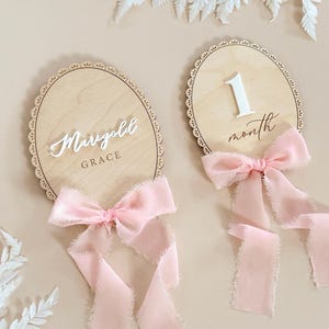 May include: Two oval wooden plaques with scalloped edges, each adorned with a light pink frayed ribbon bow. One plaque features the white 3D text "Marigold GRACE," while the other displays the white 3D number "1" above the script text "month."