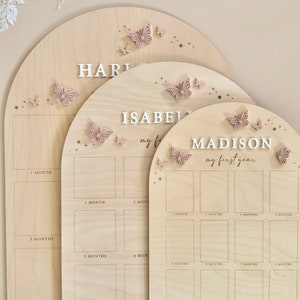 Puede incluir: Tres tablas de crecimiento de madera con un acabado de madera clara y detalles de mariposas rosas. Las tablas est&aacute;n etiquetadas como "Hart", "Isabel" y "Madison". Cada tabla tiene un espacio para una foto para cada mes del primer a&ntilde;o.