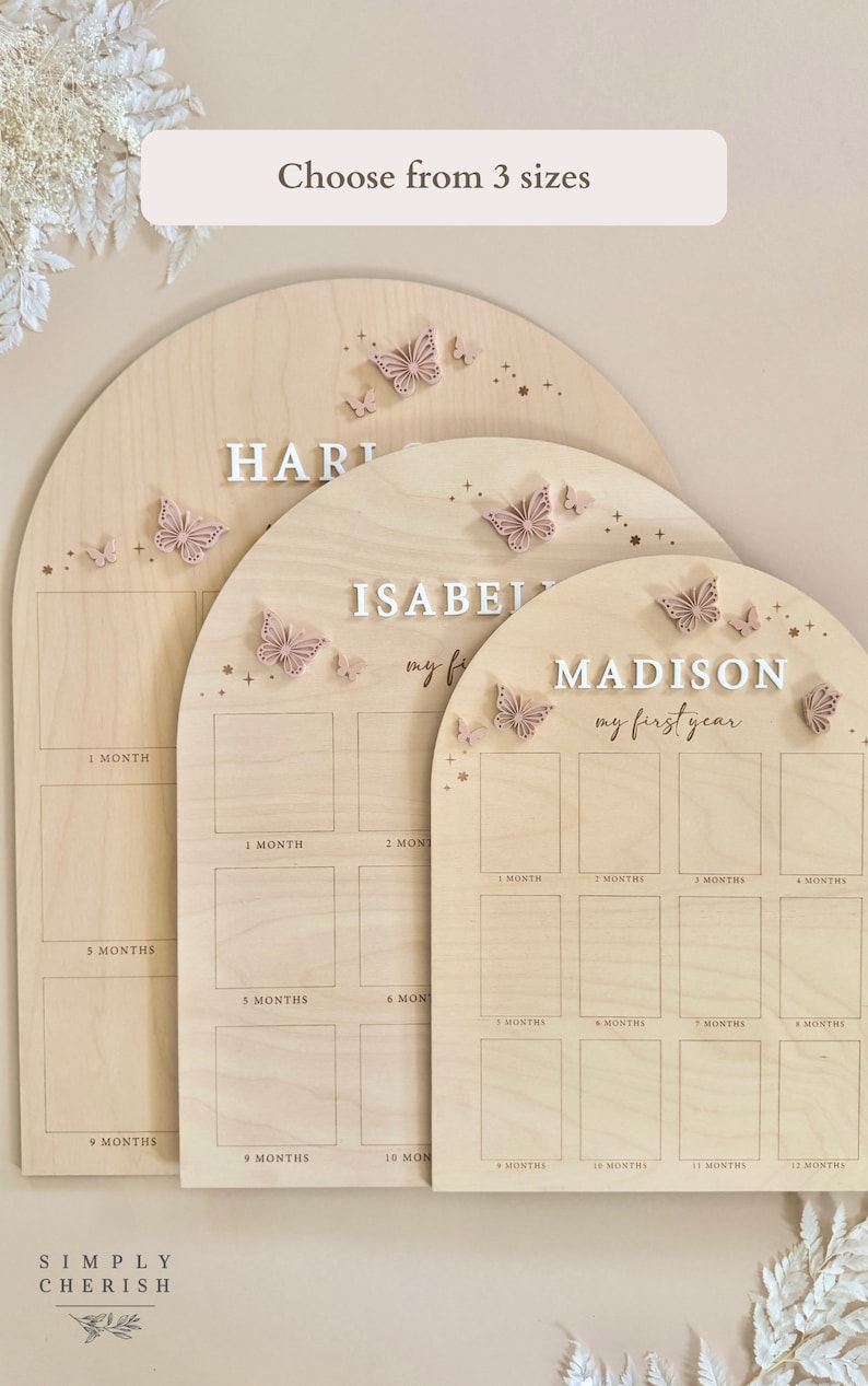 Puede incluir: Tres tablas de crecimiento de madera con un acabado de madera clara y detalles de mariposas rosas. Las tablas est&aacute;n etiquetadas como "Hart", "Isabel" y "Madison". Cada tabla tiene un espacio para una foto para cada mes del primer a&ntilde;o.