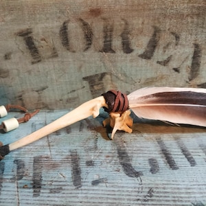 Puede incluir: Una herramienta ceremonial artesanal con una gran pluma con marcas marrones y blancas, unida a un mango de hueso con detalles de cuero y hueso. La herramienta descansa sobre una superficie de madera azul desgastada.