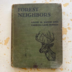May include: An antique book titled "Forest Neighbors" with a faded green cover. The cover features black text and a detailed illustration of a deer's head. The book's title and author's names are visible.