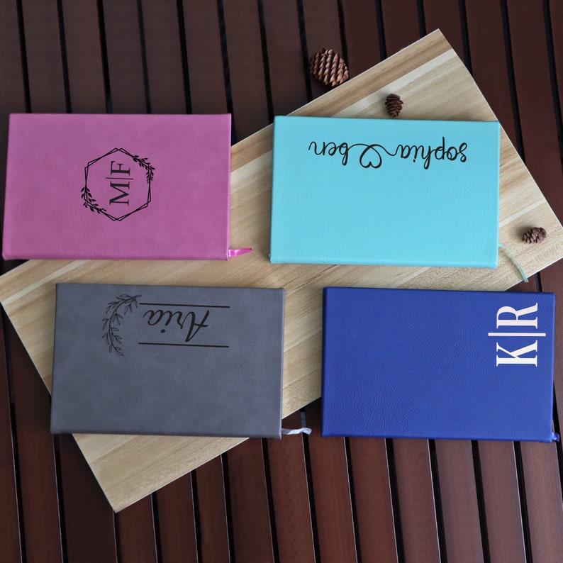 May include: Four personalized notebooks with different colors and designs. The notebooks are on a wooden surface. The designs include a monogram, a floral design, a heart, and a name.