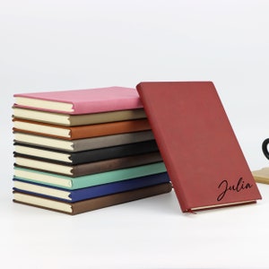May include: A stack of 12 leather-bound journals in various colors, including pink, brown, gray, black, teal, blue, and brown. The top journal is red and has the name "Julia" written on the front.