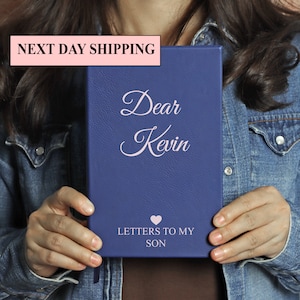 May include: A blue leather journal with the text "Dear Kevin" and "Letters to My Son" in pink lettering. A pink heart symbol is between the two lines of text.