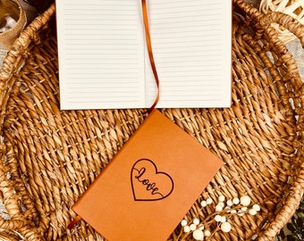 May include: A brown leather journal with a heart and the word "love" inscribed on the front cover. The journal is open to reveal lined pages and is sitting on a woven basket.
