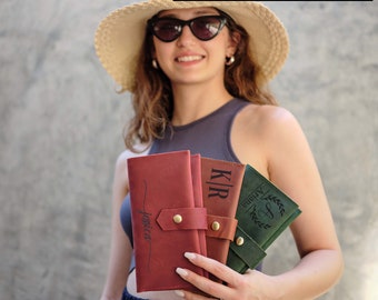 May include: Three personalized leather wallets in different colors. The wallets are held by a person wearing a straw hat and sunglasses. The wallets are engraved with the names "Jessica", "KIR", and "A".