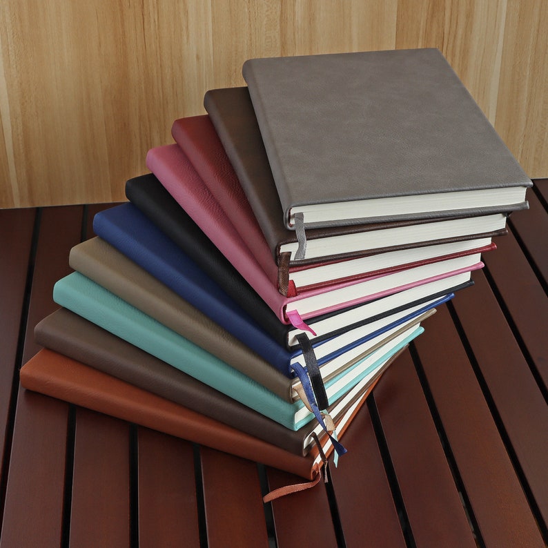 May include: A stack of ten leather-bound journals in various colours, including brown, teal, grey, pink, and blue. The journals are arranged in a fan shape, with the top journal being grey.