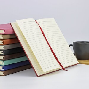 May include: A stack of notebooks with different coloured covers, including pink, brown, grey, blue, and green. The top notebook is open, revealing lined pages and a red ribbon bookmark.