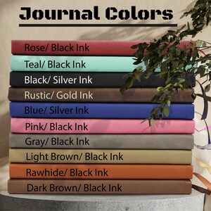 May include: A stack of ten leather-bound journals in various colours, including rose, teal, black, rustic, blue, pink, grey, light brown, rawhide, and dark brown. Each journal has a different colour and ink combination, as indicated by the text on the front of each journal.