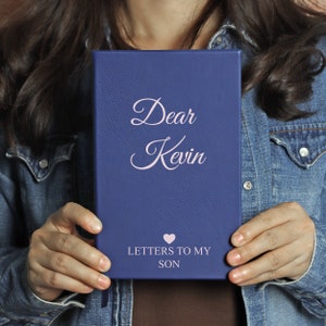 May include: A blue leather journal with the text "Dear Kevin" and "Letters to My Son" in pink lettering. A pink heart symbol is between the two lines of text.