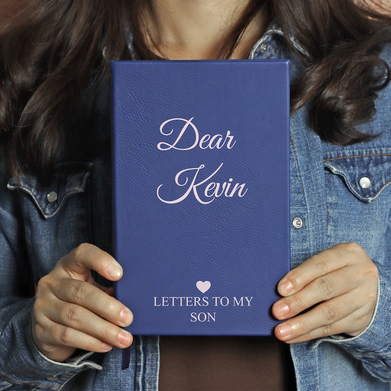 May include: A blue leather journal with the text "Dear Kevin" and "Letters to My Son" in pink lettering. A pink heart symbol is between the two lines of text.