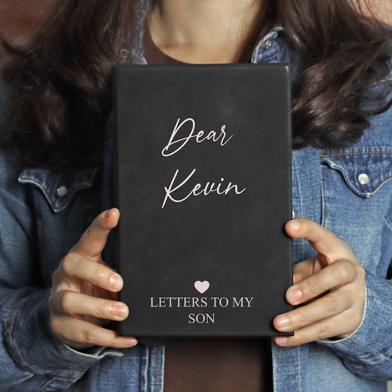 May include: Black leather journal with the text "Dear Kevin" and "Letters to My Son" in white lettering. A pink heart symbol is above the text "Letters to My Son".
