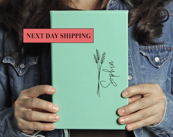 May include: A mint green leather journal with a black wheat stalk design and the name "Sophia" written in cursive.