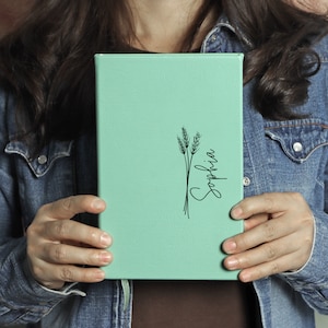 May include: A mint green leather-bound journal with a black wheat stalk design and the name "Sophia" written in cursive.