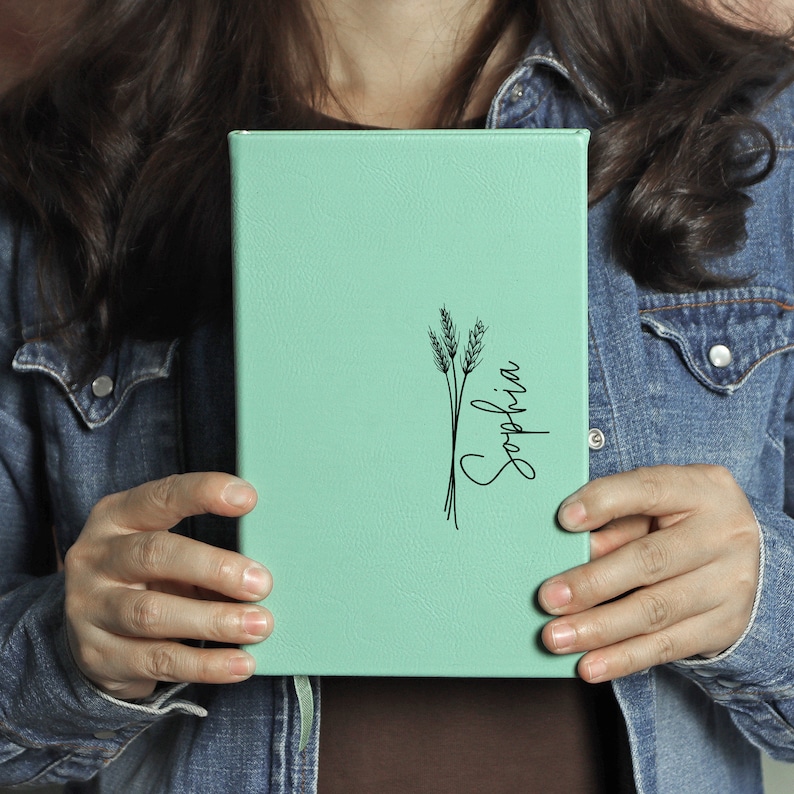 May include: A mint green leather-bound journal with a black wheat stalk design and the name "Sophia" written in cursive.