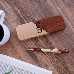 May include: A wooden pen case with a two-toned design, one side is light wood and the other is dark wood. The case has the name "Sam" engraved on it. A wooden pen with a gold trim and the name "Sam" engraved on it is next to the case.