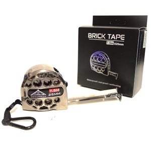 May include: A silver-colored metal measuring tape with a hollow, circular cutout design on its casing. The tape is labeled 7.5M and 25MM. It includes a black wrist strap and is displayed next to its black retail box labeled BRICK TAPE 7.5M X 25MM.