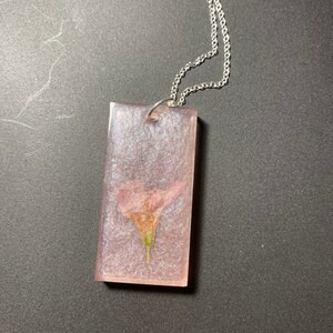 May include: A silver chain necklace with a rectangular pendant featuring a pressed pink flower encased in clear resin.