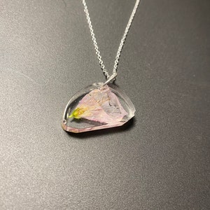 May include: A clear resin pendant necklace with a silver chain. The pendant is a unique shape, with a dried pink and yellow flower encased inside. The necklace is displayed on a dark gray surface.
