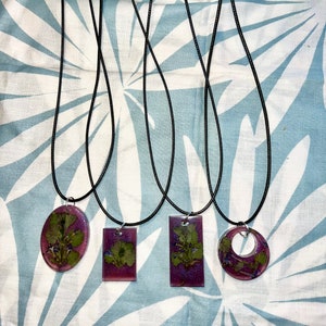 May include: Four resin pendant necklaces with black cords. Each pendant features a different shape: oval, square, and rectangle, and a circle. The pendants contain dried green leaves and purple glitter.