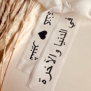 May include: Two white fabric ribbons with black Arabic calligraphy and a black heart. The ribbons are tied together with a white ribbon.