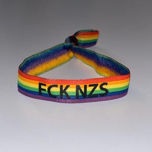 May include: A fabric wristband featuring a horizontal rainbow stripe pattern in red, orange, yellow, green, blue, and purple. The text FCK NZS is embroidered in black thread across the centre of the band. A small black plastic bead secures the ends.