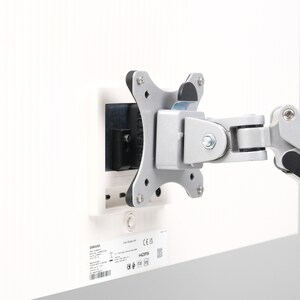 May include: A silver and black monitor arm attached to a white surface. The arm has multiple joints and a mounting plate with four screws. A black rectangular component is also attached to the white surface.