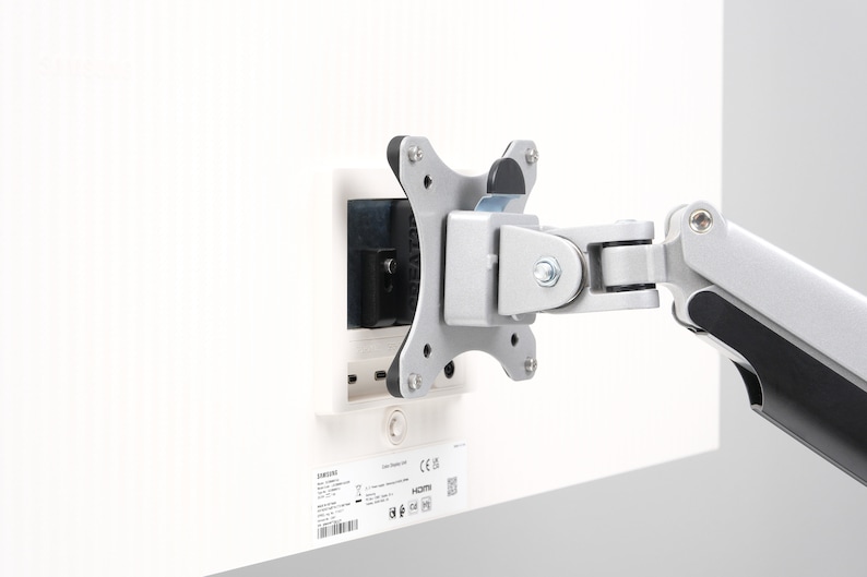 May include: A silver and black monitor arm attached to a white surface. The arm has multiple joints and a mounting plate with four screws. A black rectangular component is also attached to the white surface.