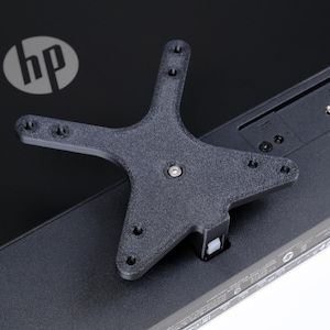Op de afbeelding: Een zwarte, stervormige beugel met meerdere schroefgaten is bevestigd aan de achterkant van een zwarte computermonitor. Het HP-logo is zichtbaar. De monitor heeft verschillende poorten en connectoren.