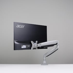 Może przedstawiać: Czarny monitor komputerowy Acer zamontowany na srebrno-szarym, regulowanym ramieniu. Monitor znajduje się na jasnoszarym tle, z widocznym logo Acer. Ramię jest przymocowane do białej powierzchni.