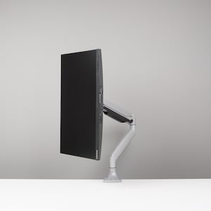 May include: A black computer monitor mounted on a silver and grey adjustable arm. The monitor is positioned to the side, displaying a blank black screen. The arm is attached to a white surface.