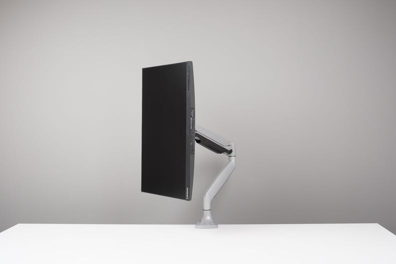 May include: A black computer monitor mounted on a silver and grey adjustable arm. The monitor is positioned to the side, displaying a blank black screen. The arm is attached to a white surface.