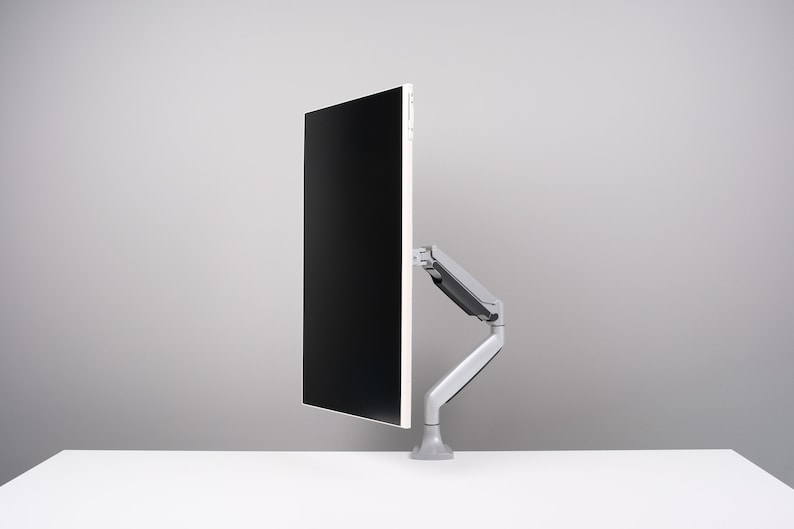 May include: A modern computer monitor with a black screen is mounted on a gray, adjustable arm. The arm is attached to a gray base, and the setup is on a white surface against a gray background. The monitor is oriented vertically.