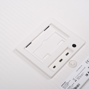 May include: A white Samsung Color Display Unit with a square recessed area, ports, and a small circular button. The unit has a label with text and a QR code. The background is white.