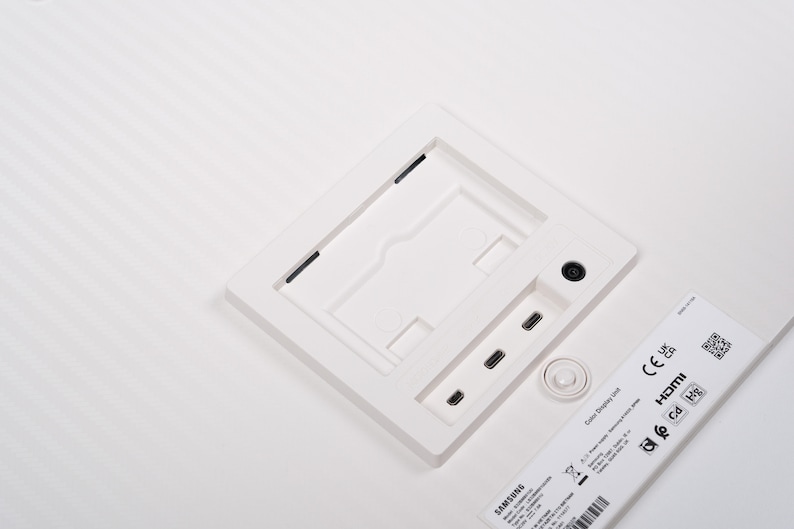 May include: A white Samsung Color Display Unit with a square recessed area, ports, and a small circular button. The unit has a label with text and a QR code. The background is white.